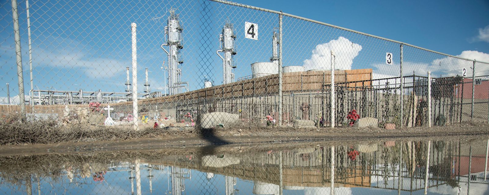 Graves with crosses and flowers beside a fenced industrial gas facility, reflected in floodwater